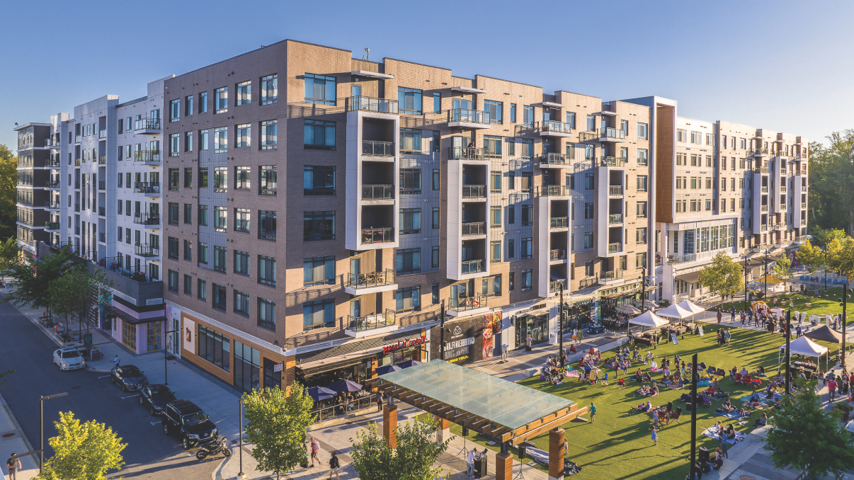 A mixed-use urban building in Merriweather, Maryland, with shops and apartments overlooks a vibrant park where people gather on a sunny day.