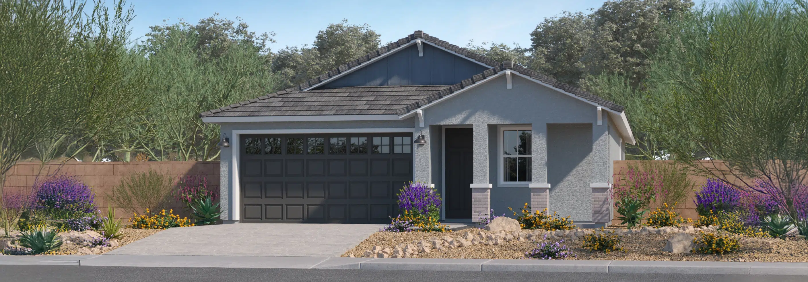 Single-story house with a gray facade and dark garage door, surrounded by desert landscaping with rocks and flowering plants, and trees in the background.