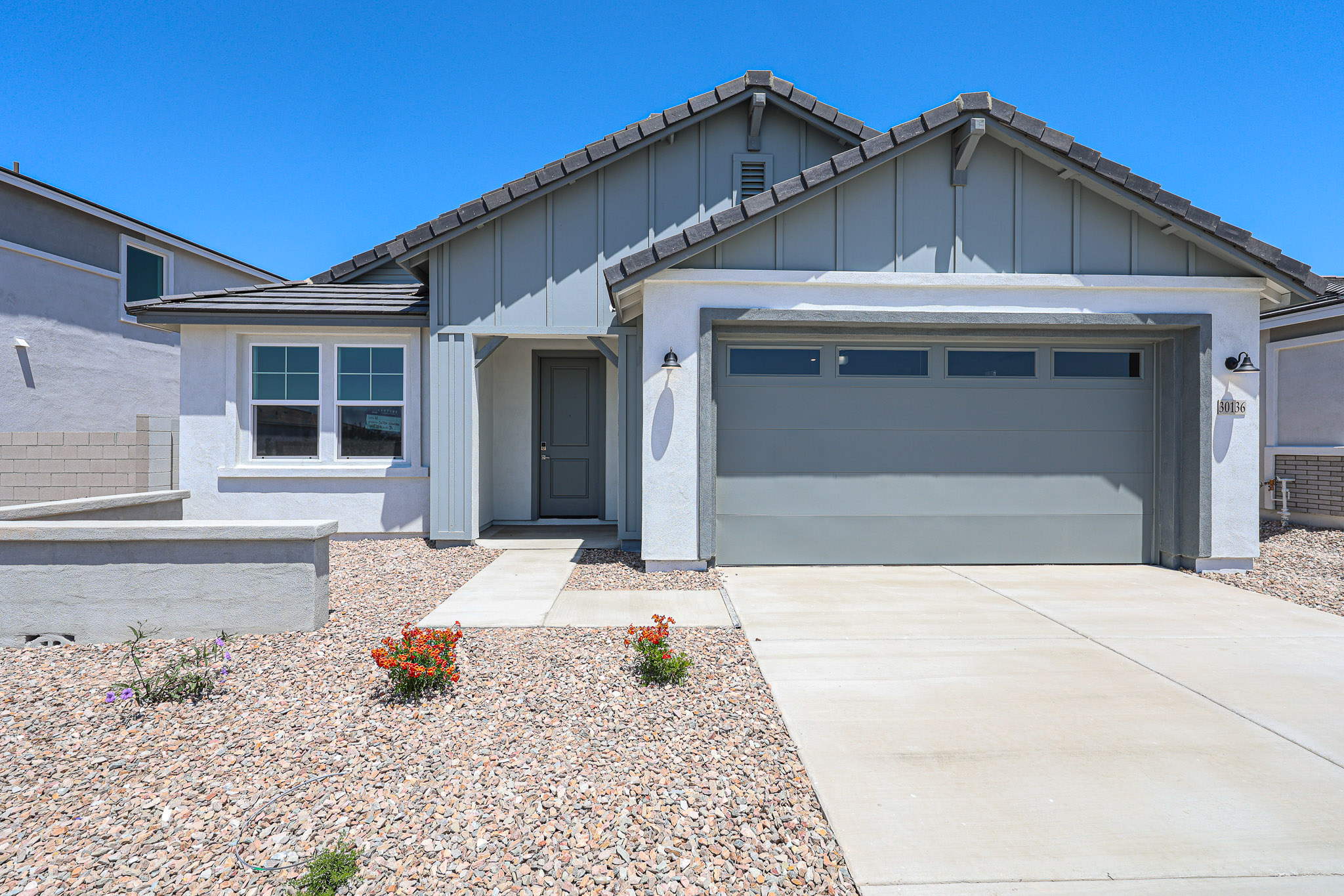 Single-story house with gray siding, a two-car garage, small front porch, and low-maintenance gravel landscaping with a few flowering plants under a clear blue sky.