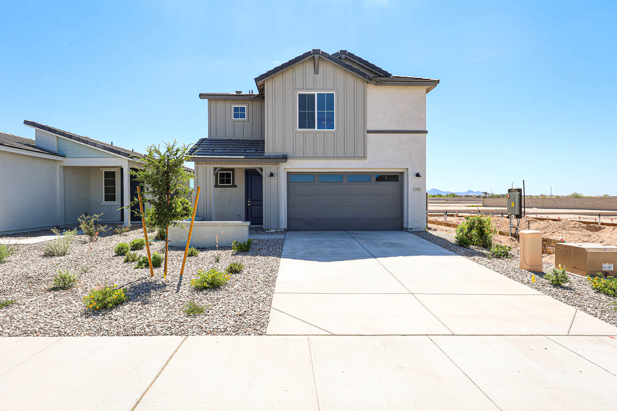 Two-story modern house with a gray garage door, landscaped front yard with rocks and plants, and a concrete driveway under a clear blue sky.