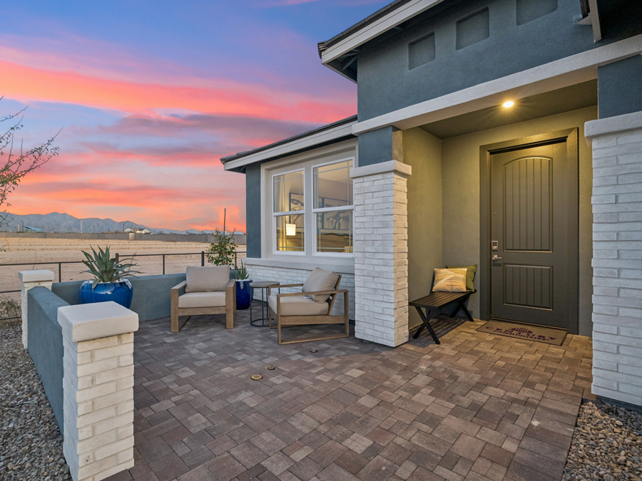 Modern house with a covered front porch, two chairs, a small table, potted plants, and a brick walkway at sunset.