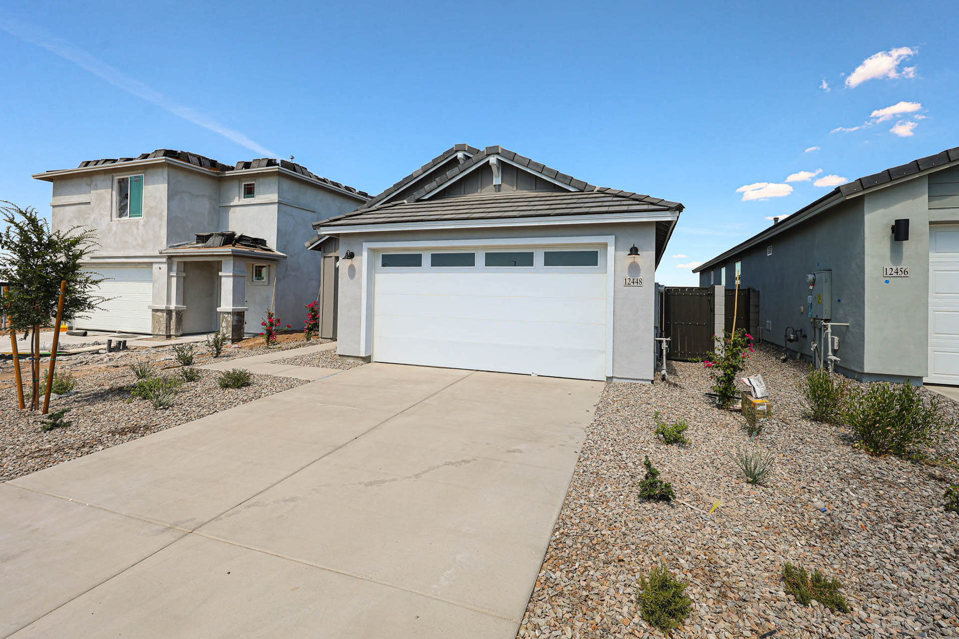 Single-story house with a light gray exterior, double garage, concrete driveway, and minimalist desert landscaping under a clear sky.