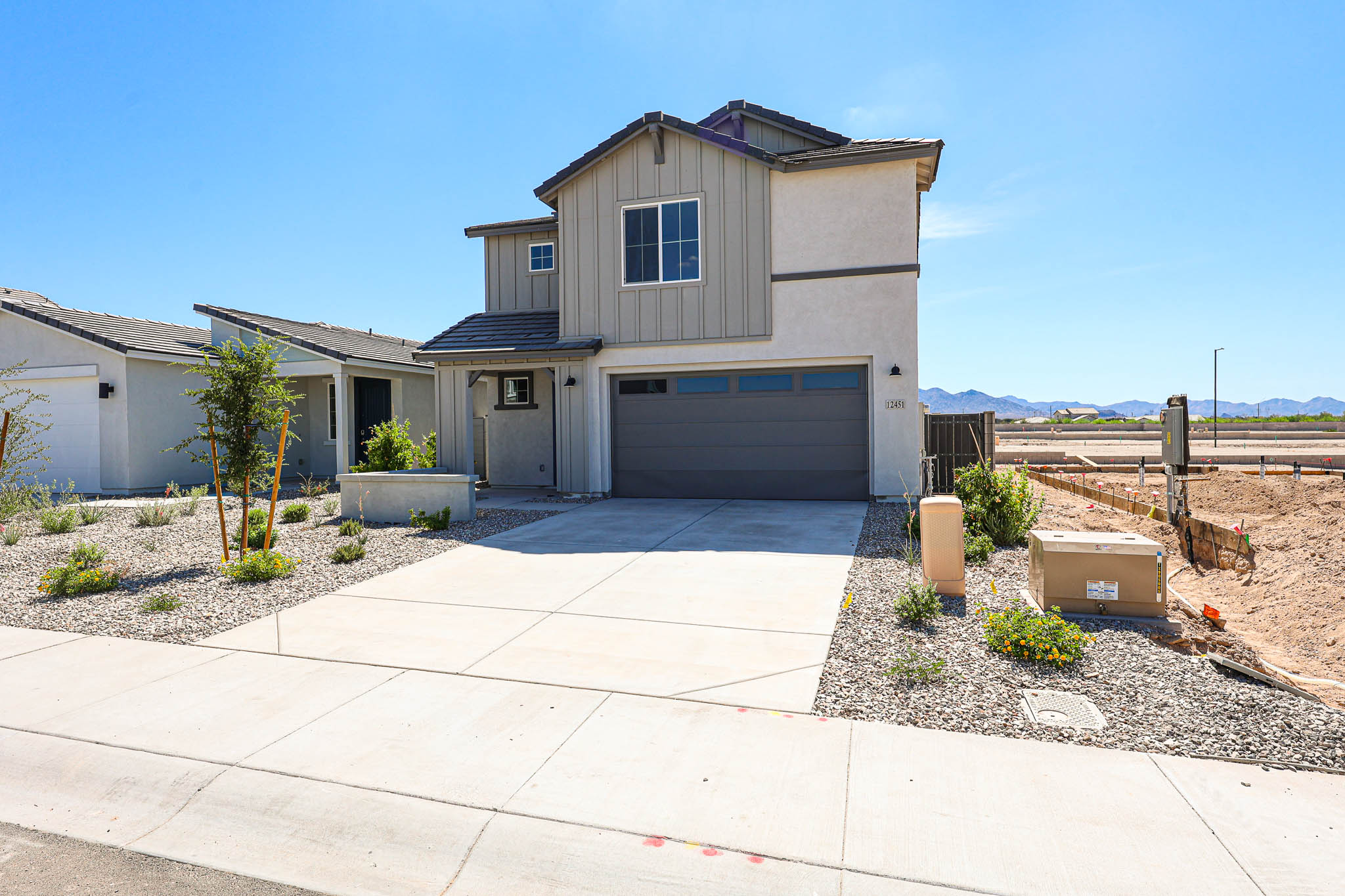 Two-story modern house with gray garage door, small front yard with minimal landscaping, and a wide driveway under a clear blue sky. Construction is visible to the right.