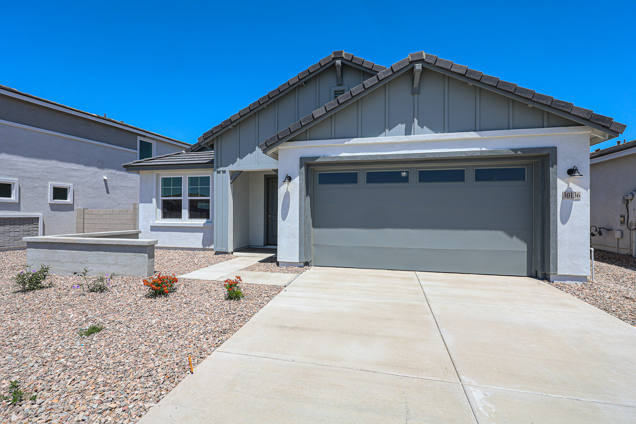 Single-story modern house with a two-car garage, gray roof, and light exterior, surrounded by a gravel yard and minimal landscaping under a clear blue sky.