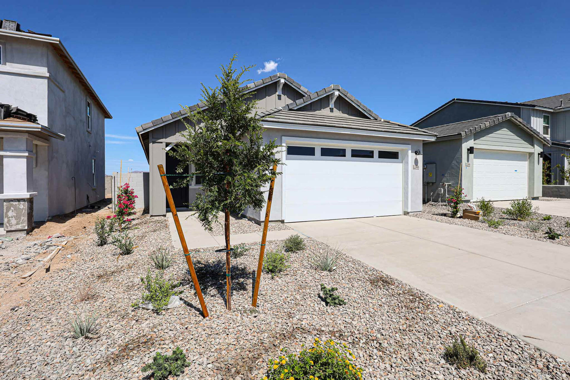 Single-story house with a white garage door, gravel yard landscaping, young tree in front, and concrete driveway under a clear blue sky.