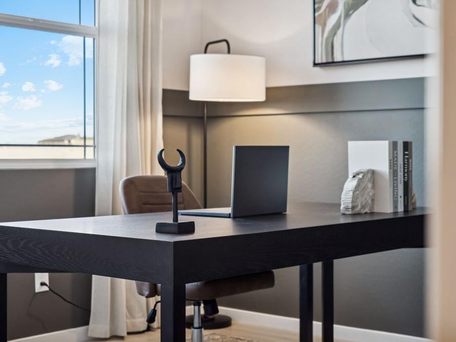 Modern home office with a black desk, closed laptop, decorative items, a brown chair, lamp, and window with white curtains showing a blue sky.