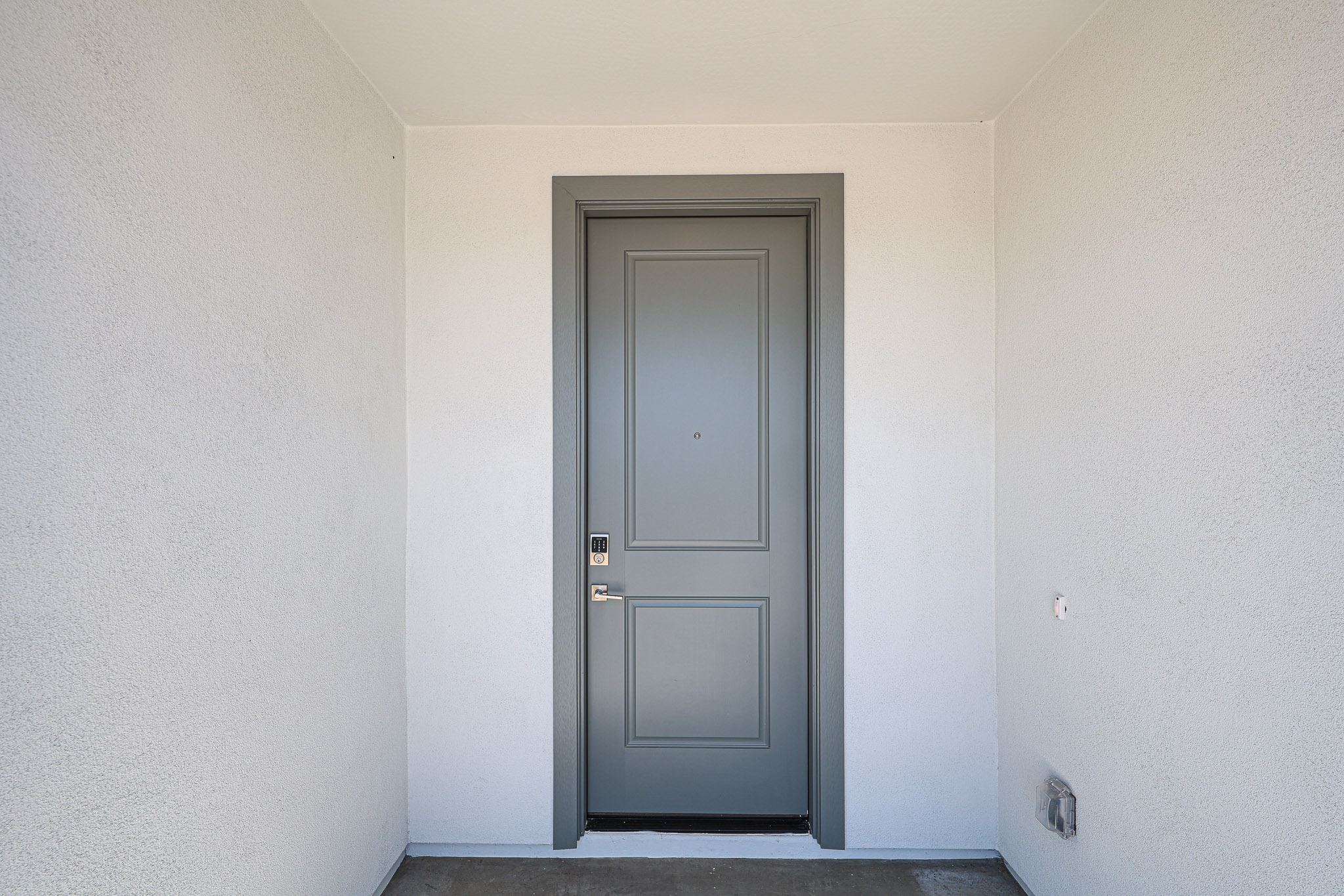 Gray front door with a rectangular design set in a white stucco wall, featuring a silver handle and digital lock, photographed from straight on.