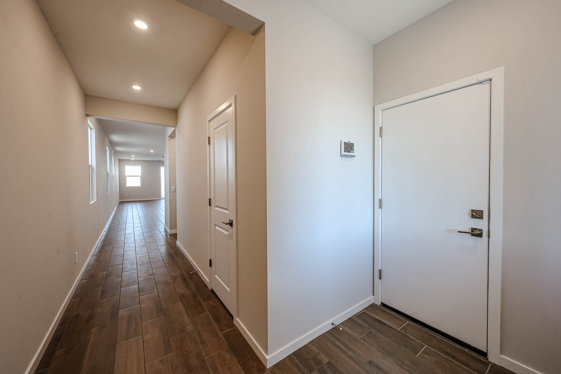 A hallway with wood-look tile flooring, white walls, a closed white door, and recessed ceiling lights leading to a bright room with windows.