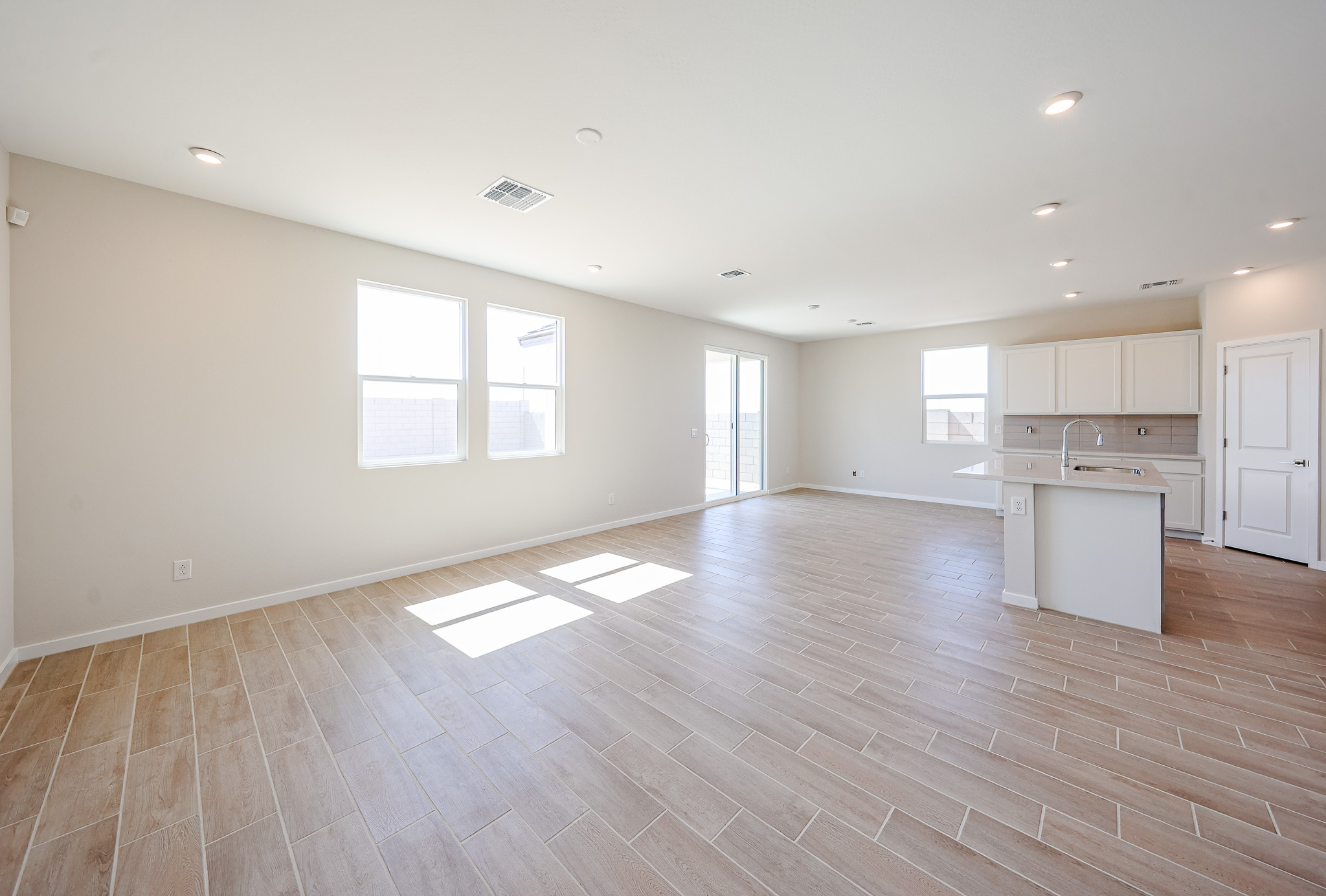 Spacious empty room with light wood tile flooring, white walls, large windows, sliding glass door, and an open kitchen with white cabinets and an island.