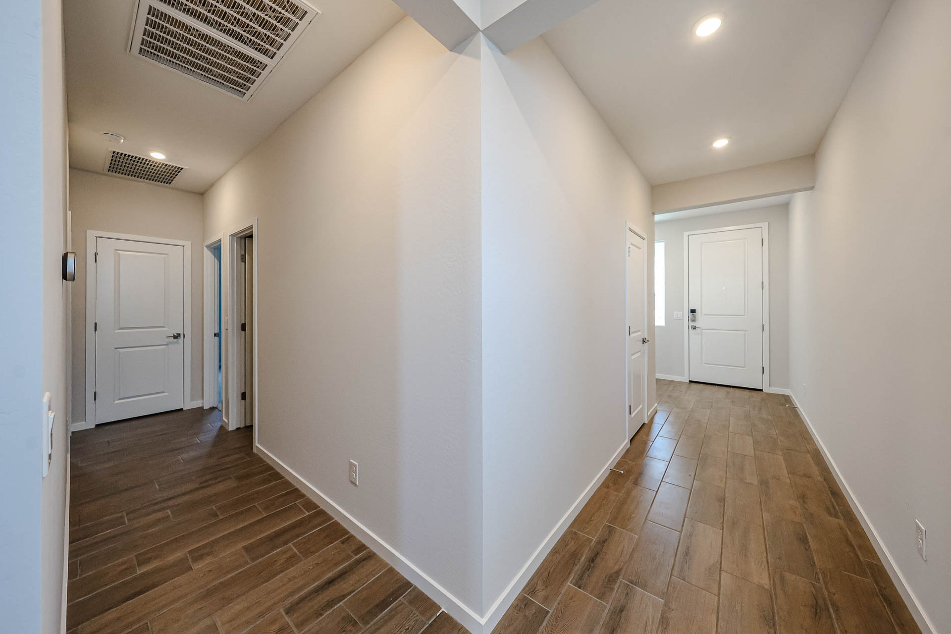 A hallway with wood-look tile flooring and white walls, splitting in two directions, each leading to several closed white doors under recessed lighting.