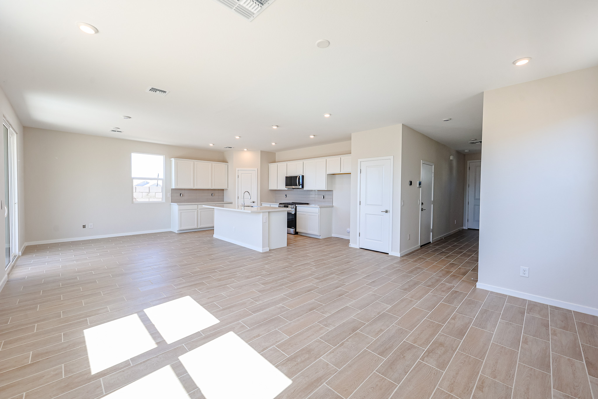 Spacious, empty open-plan living area with light wood tile flooring, white kitchen cabinets, an island, and recessed lighting. Large windows let in natural light.
