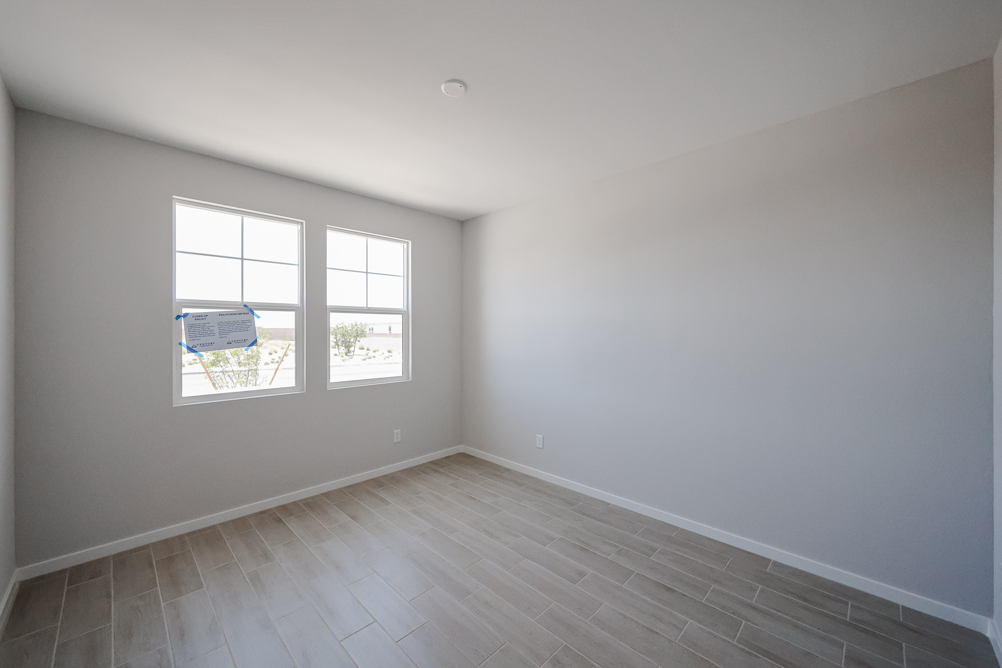 Empty room with light gray walls, two windows, tan wood-look tile flooring, and a ceiling light; sunlight is coming through the windows.