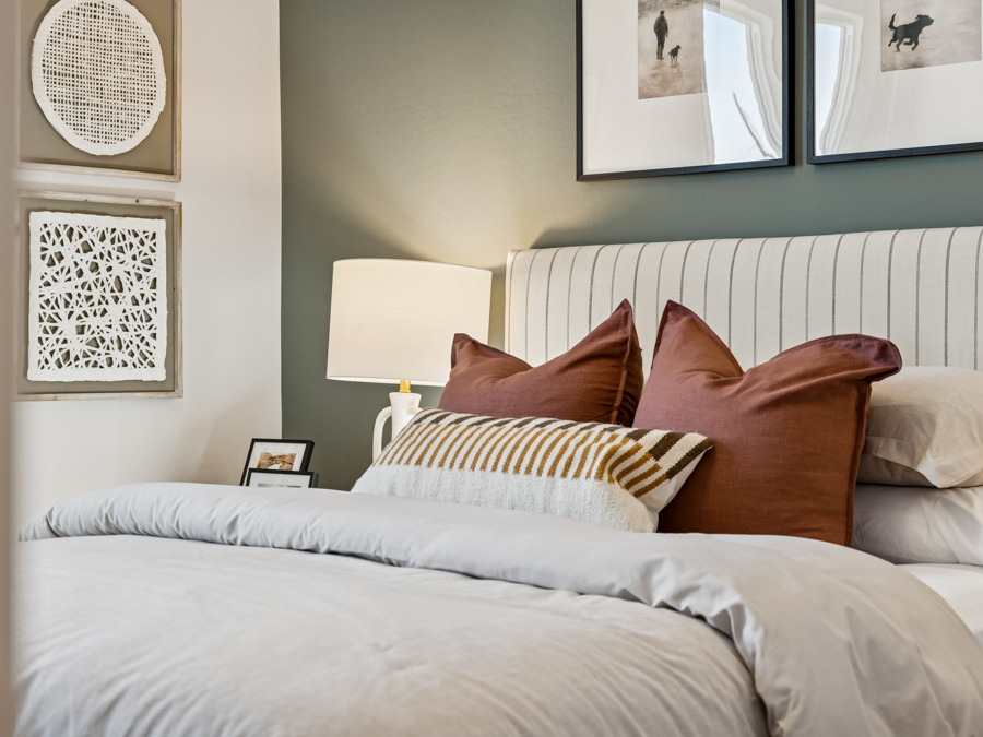 A neatly made bed with brown and striped pillows, white bedding, a lit lamp, and framed wall art in a modern bedroom.
