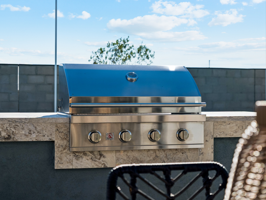 Stainless steel outdoor grill with four control knobs, built into a stone countertop on a patio under a blue sky with clouds.