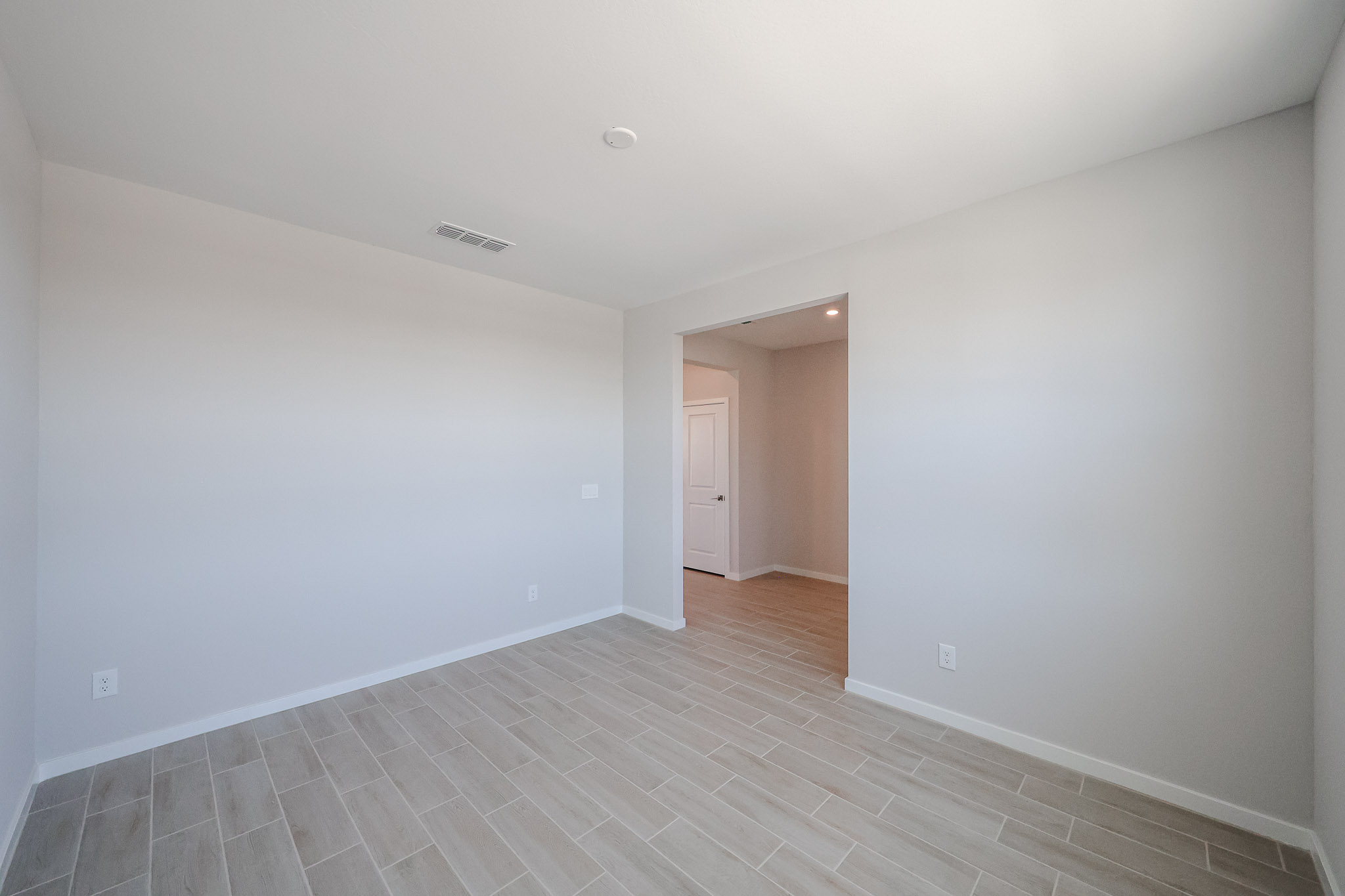 Empty room with light gray walls, light wood-look tile flooring, ceiling vent, and an open doorway leading to a hallway with a closed white door. Natural light enters from the right.