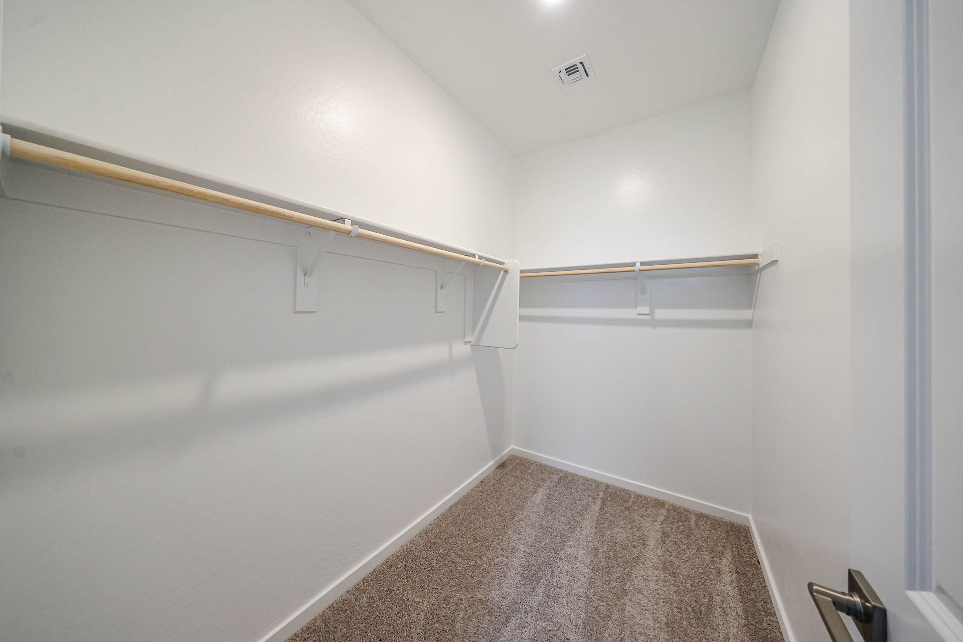Empty walk-in closet with beige carpet, white walls, and two wooden hanging rods attached to both sides of the room.