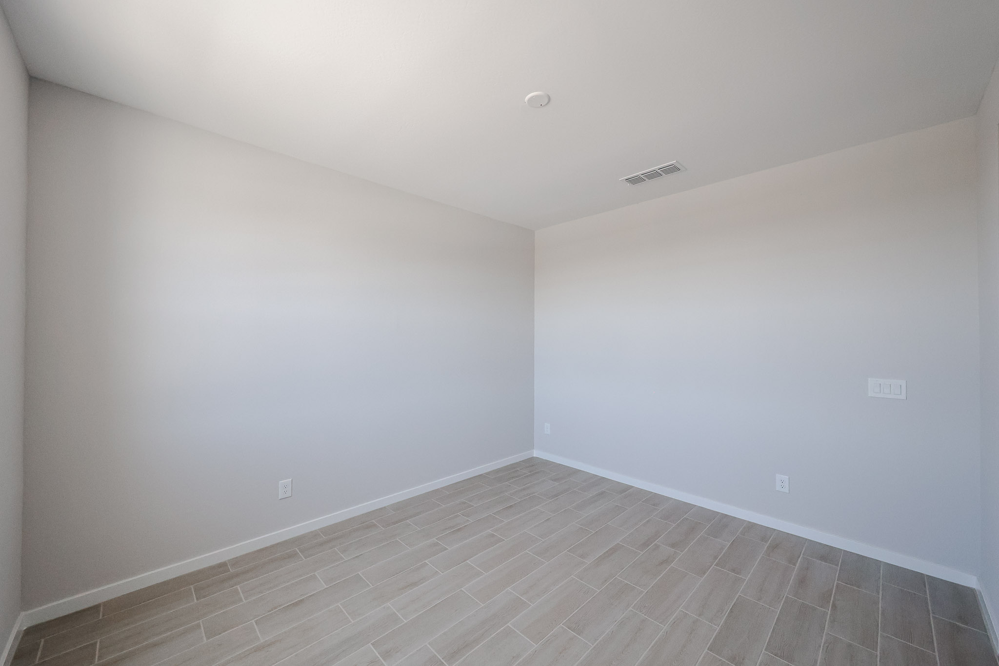 Empty room with light-colored walls, beige tile flooring, ceiling vent, and electrical outlets visible; no furniture or decor present.