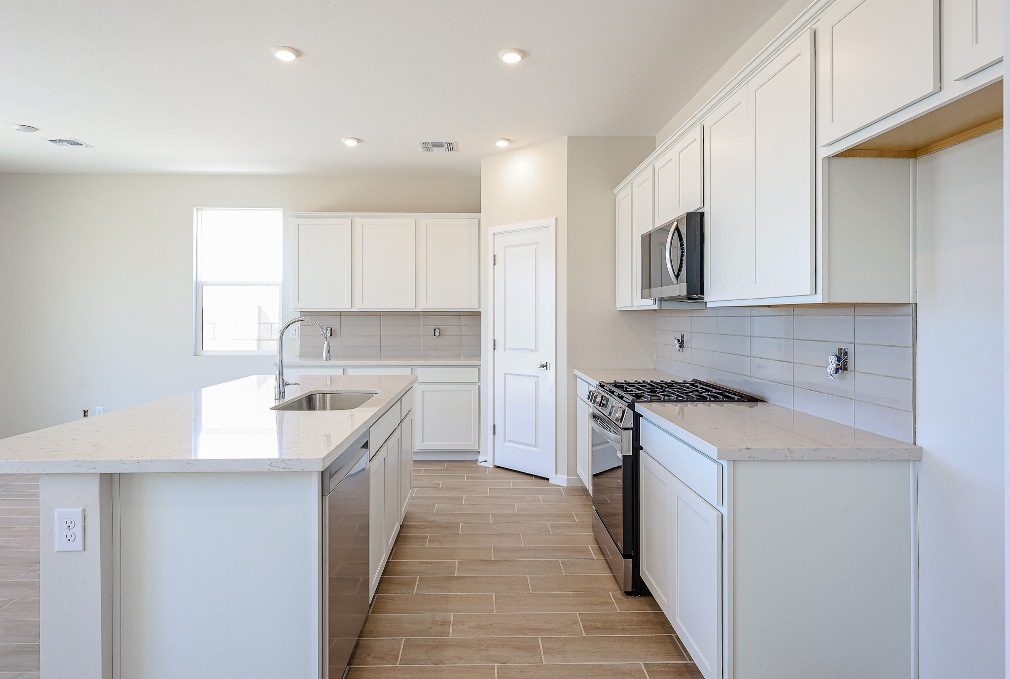 Modern kitchen with white cabinets, stainless steel appliances, a large island with sink, tile backsplash, and light wood-look tile flooring. Bright and spacious with natural light.