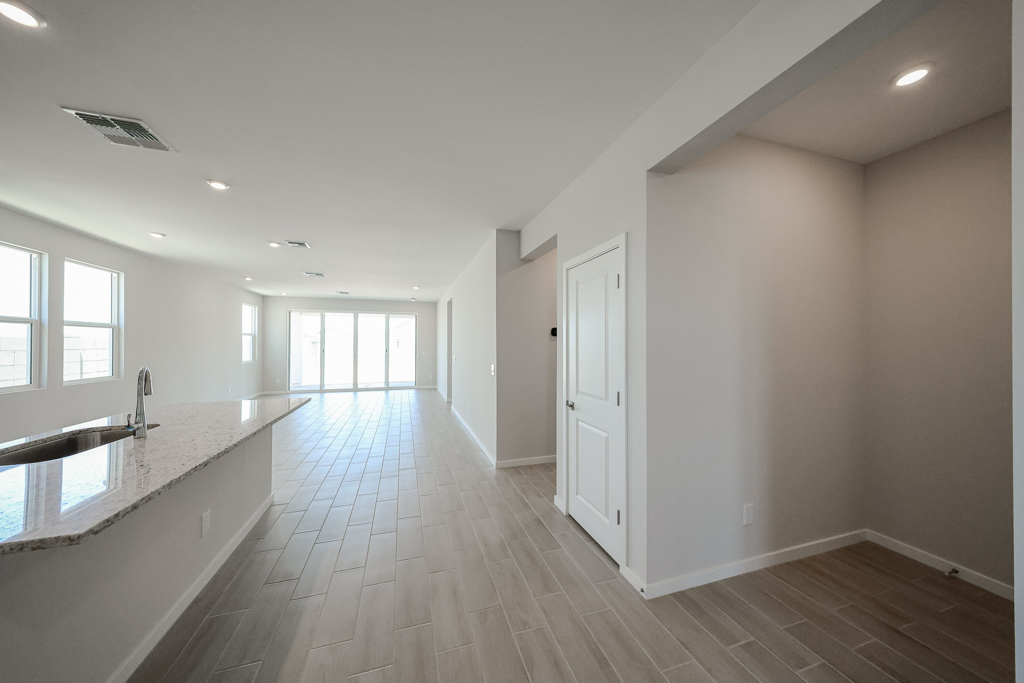 Modern, empty open-concept living space with light tile flooring, white walls, large windows, a kitchen island, and a hallway leading to other rooms.