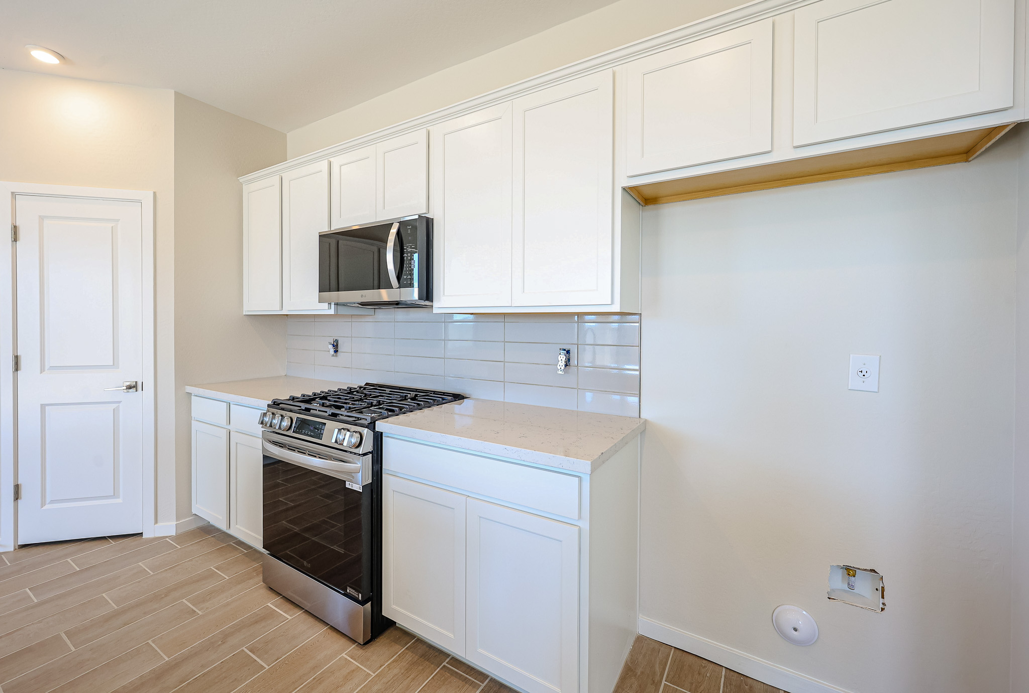 Modern kitchen with white cabinets, a stainless steel gas stove, microwave, beige tile floor, and an empty space for a refrigerator with visible power and water connections.