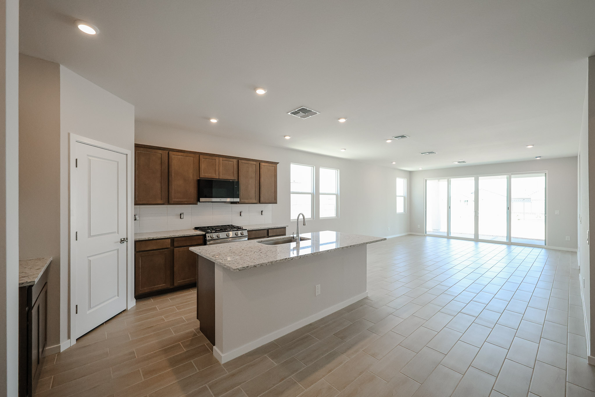 Spacious modern kitchen with brown cabinets, granite island, and open floor plan leading to a bright living area with large windows and sliding glass doors.