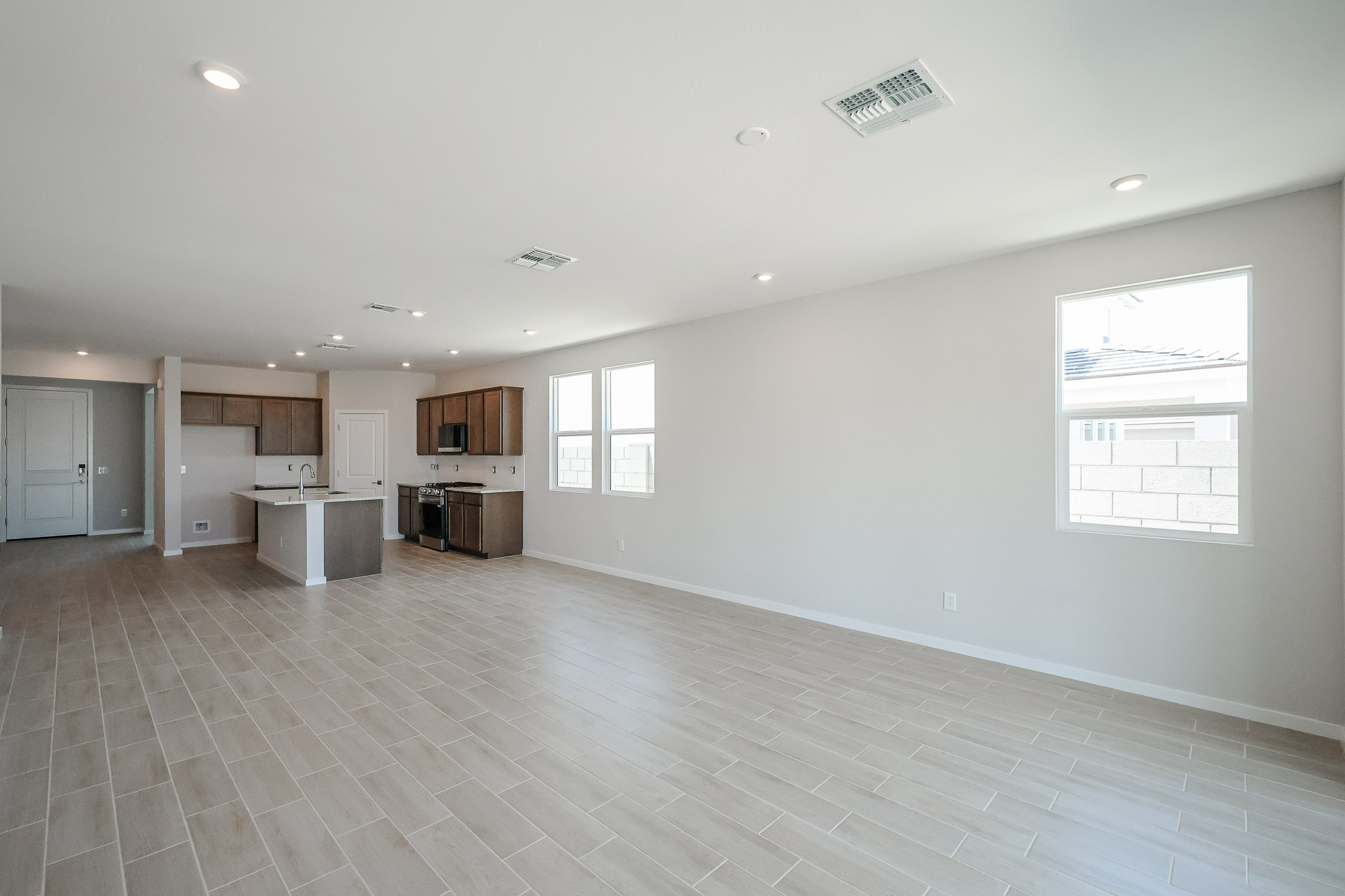Spacious, unfurnished open-concept living area with tile flooring, recessed lighting, two windows, and a kitchen with wooden cabinets and stainless steel appliances in the background.