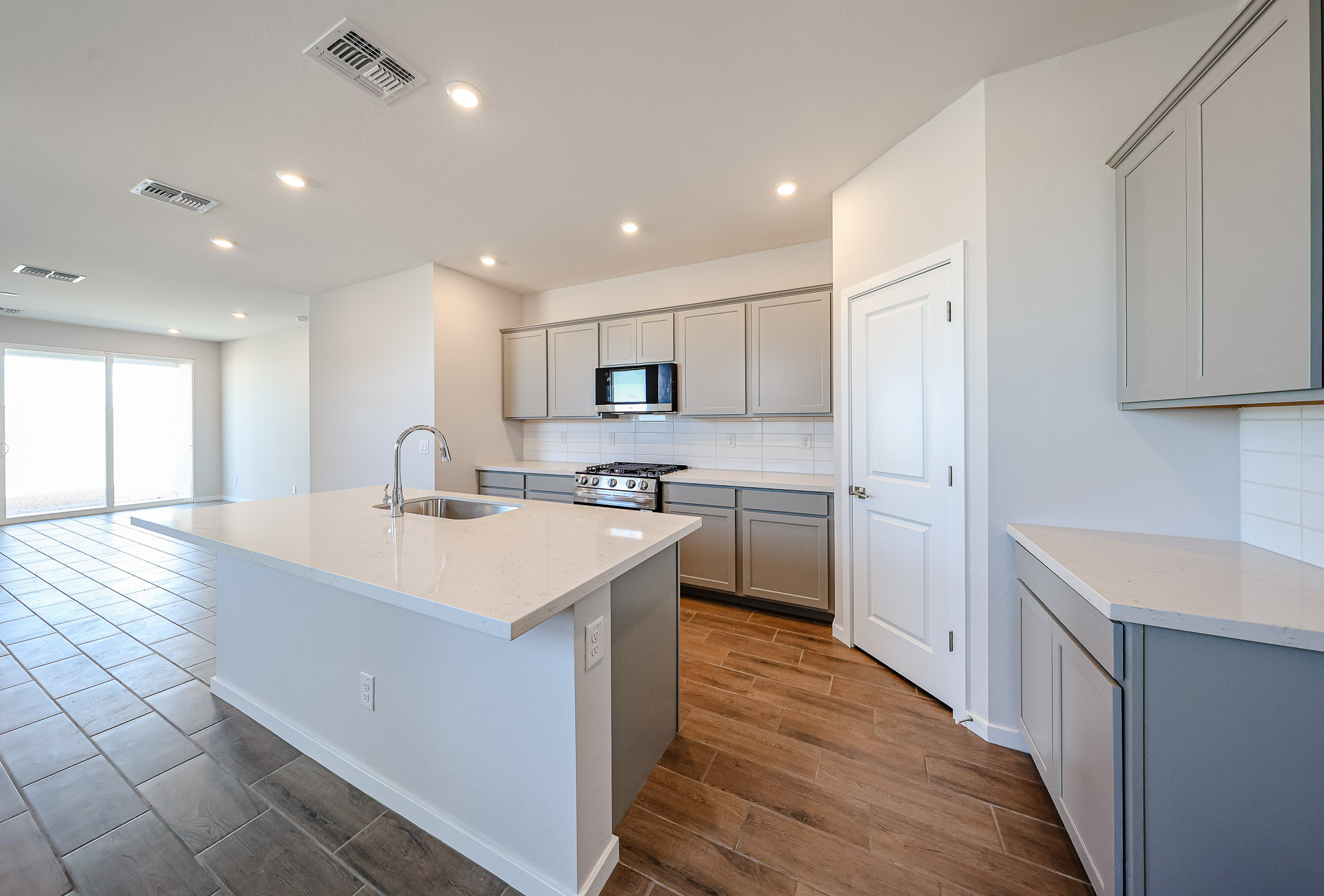 Modern kitchen with gray cabinets, stainless steel appliances, white countertops, an island with a sink, and an open layout leading to a living area with tile flooring.