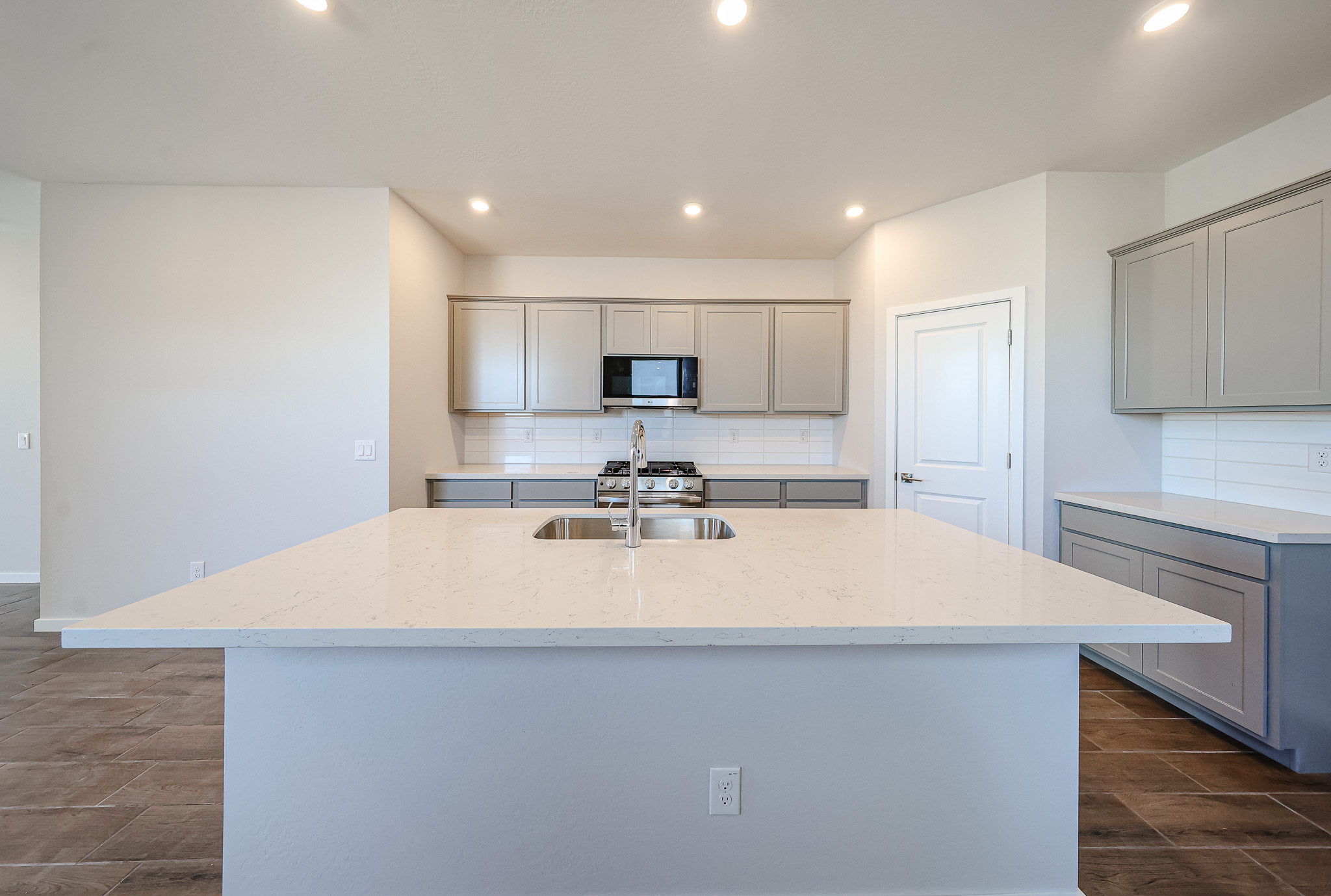 Modern kitchen with a large white island, built-in sink, gray cabinets, stainless steel appliances, and recessed lighting.