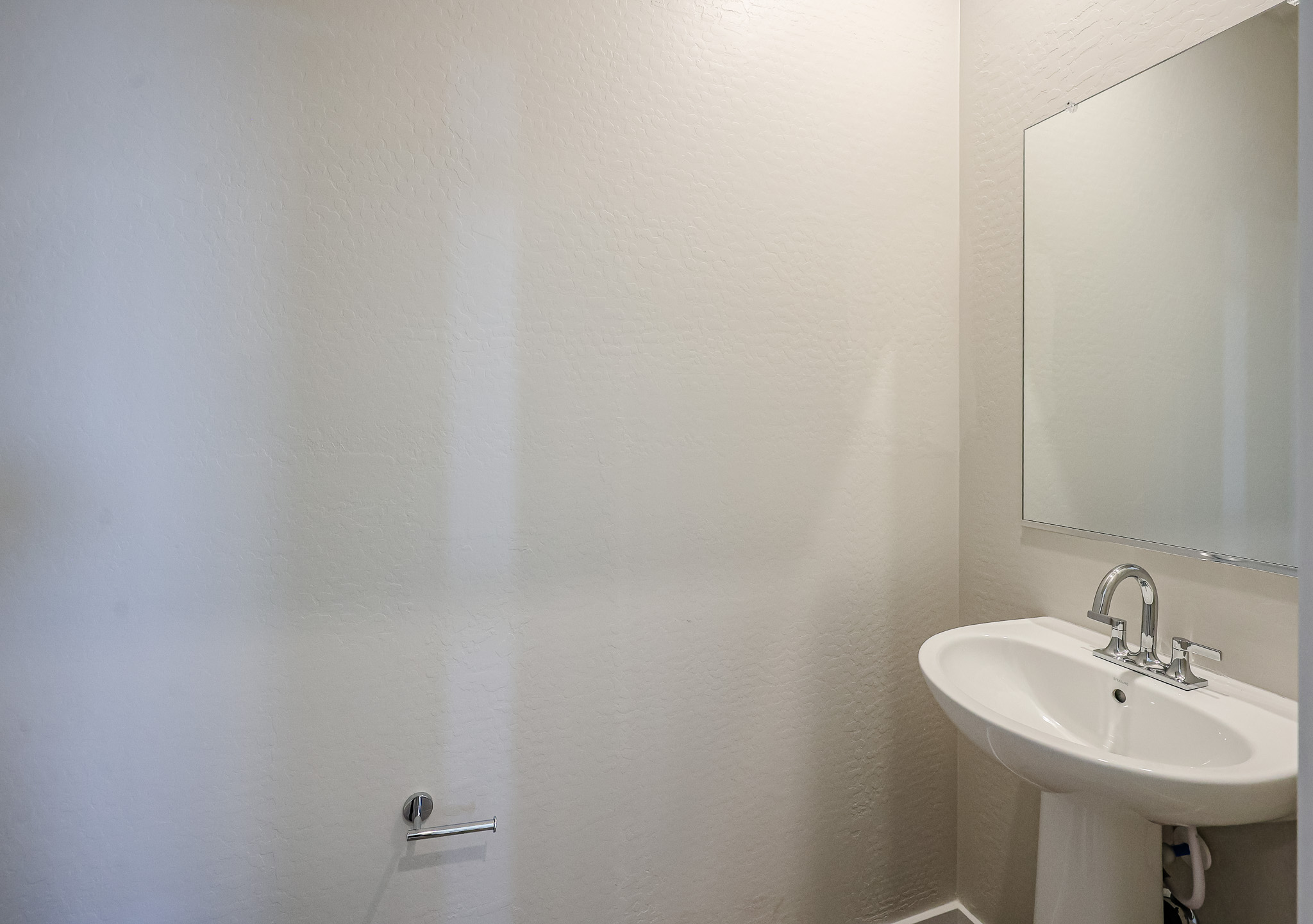Small bathroom with a white pedestal sink, chrome faucet, rectangular mirror, and empty toilet paper holder on a textured light-colored wall.