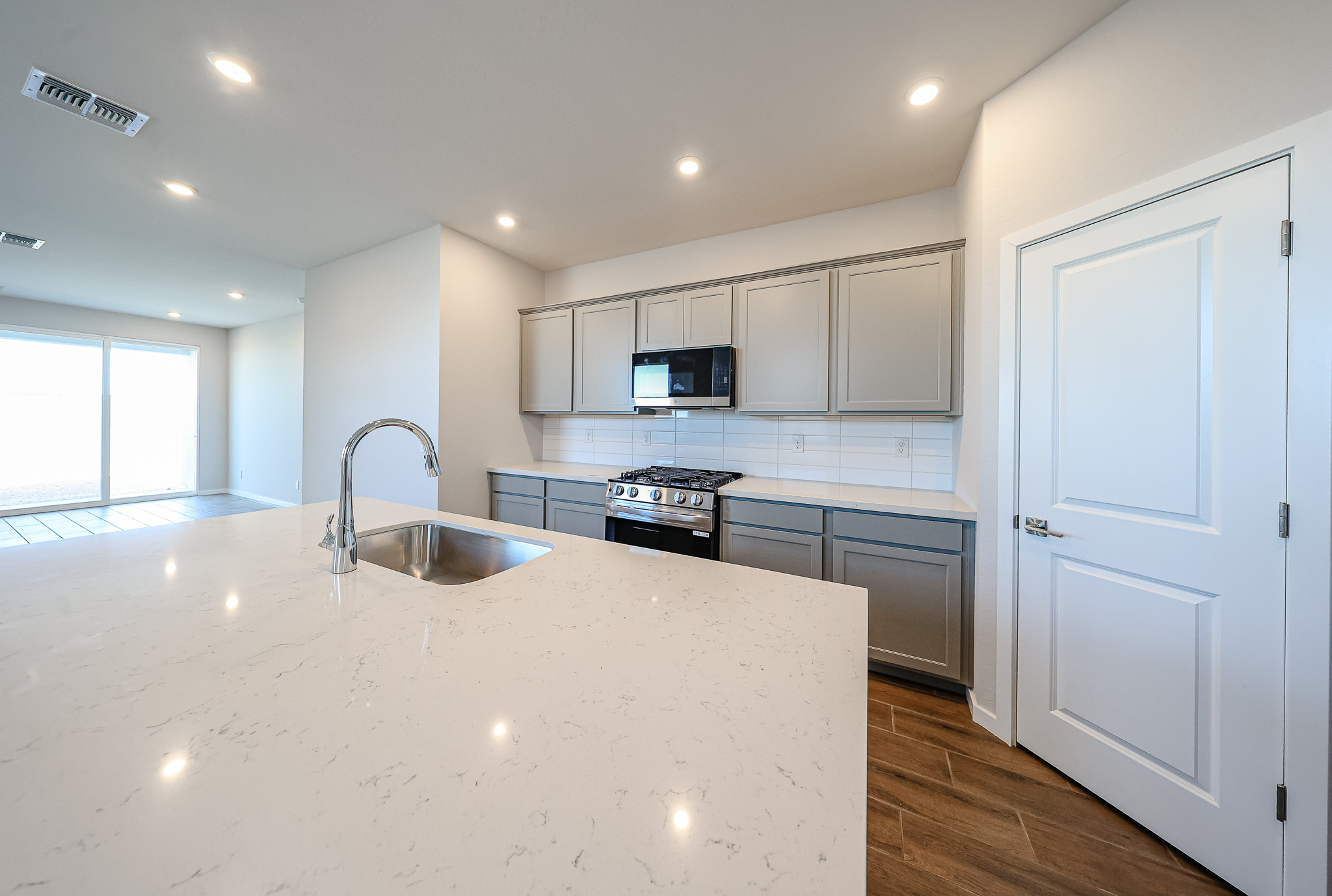 Modern kitchen with gray cabinets, stainless steel appliances, a white marble island with sink, wood flooring, and recessed lighting. Door and large window visible in the background.