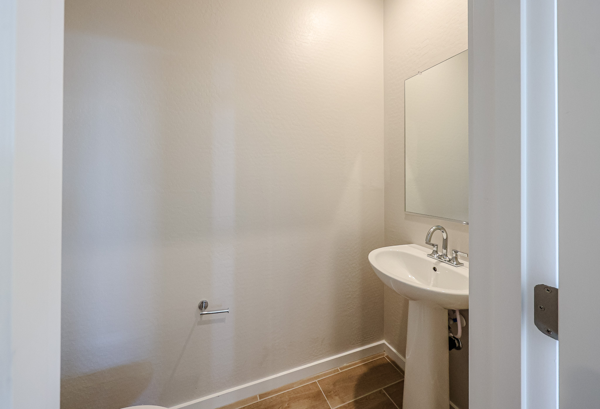Small bathroom with beige walls, pedestal sink, rectangular mirror, and an empty toilet paper holder, viewed from the doorway.