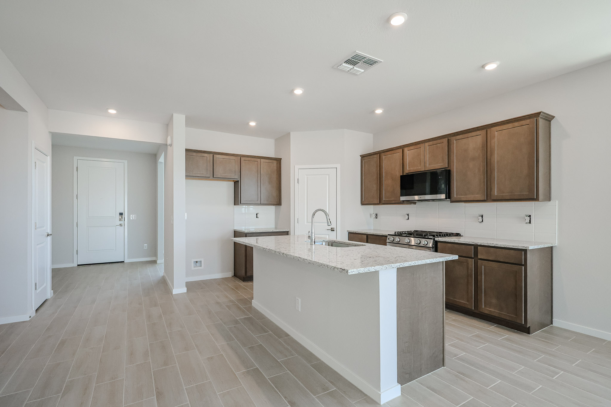 Modern kitchen with brown cabinets, white countertops, an island with a sink, built-in appliances, and light wood-look tile flooring. White walls and recessed ceiling lights throughout.