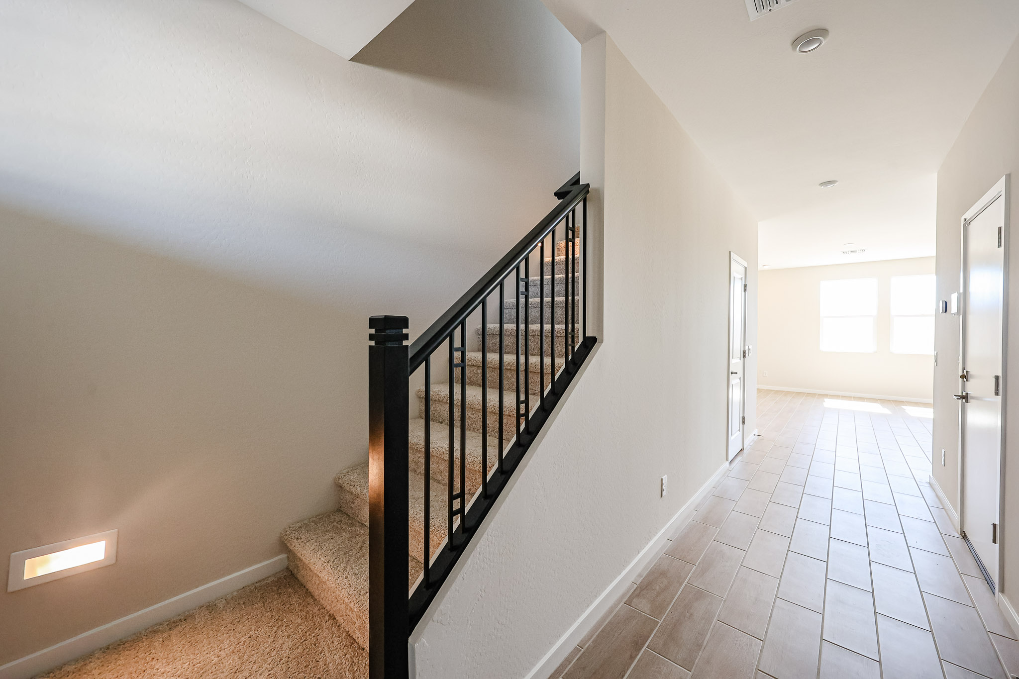 A carpeted staircase with a black railing is on the left, and a hallway with tiled flooring and white walls leads to a bright room at the end.