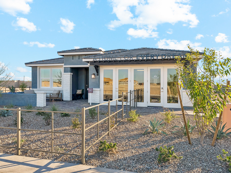 A modern, single-story house with large windows, a front porch, and desert landscaping under a blue sky with clouds.