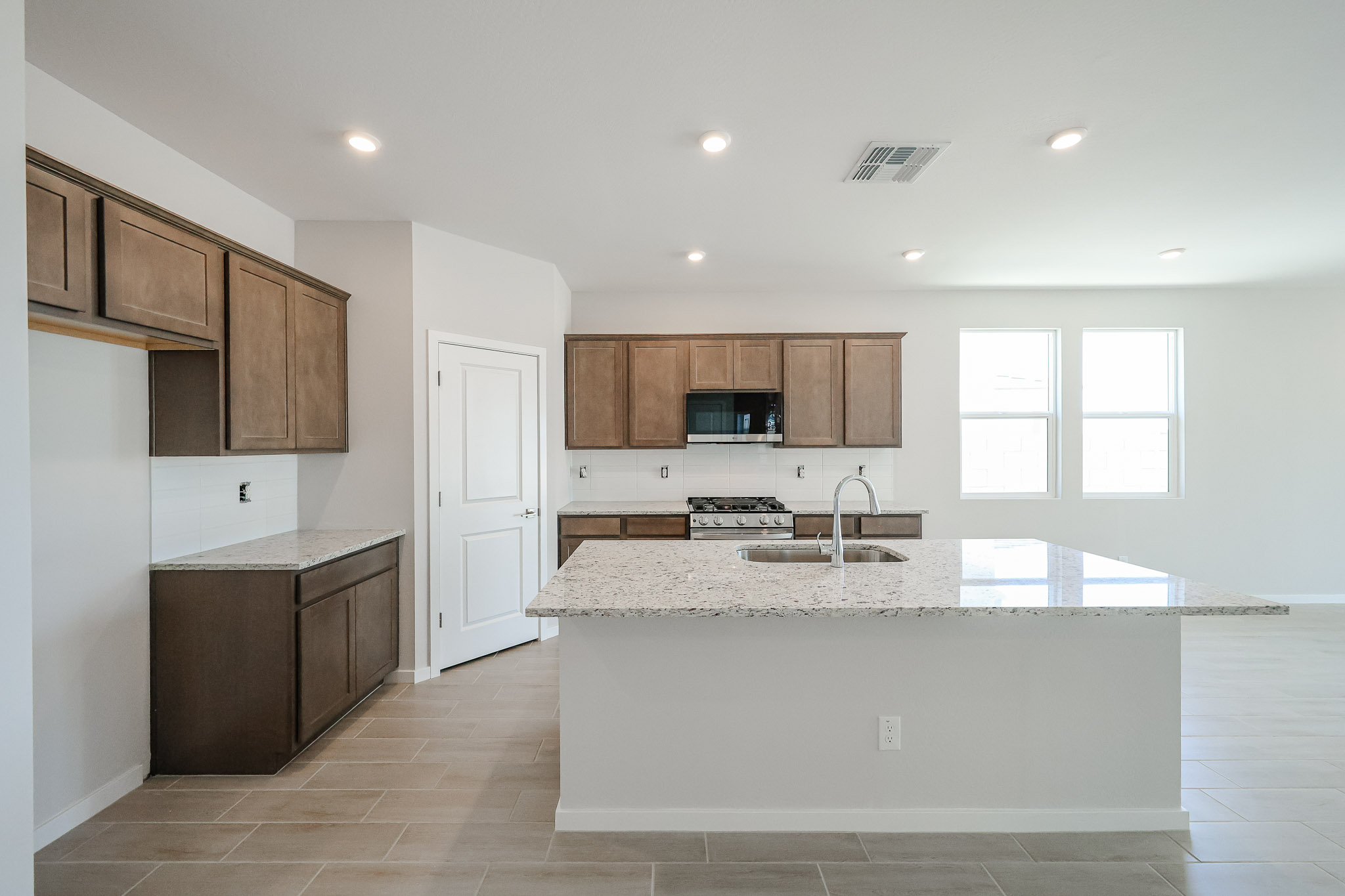 Modern kitchen with brown cabinets, granite island with sink, stainless steel appliances, and tile flooring. Two windows let in natural light. Walls and ceiling are white.