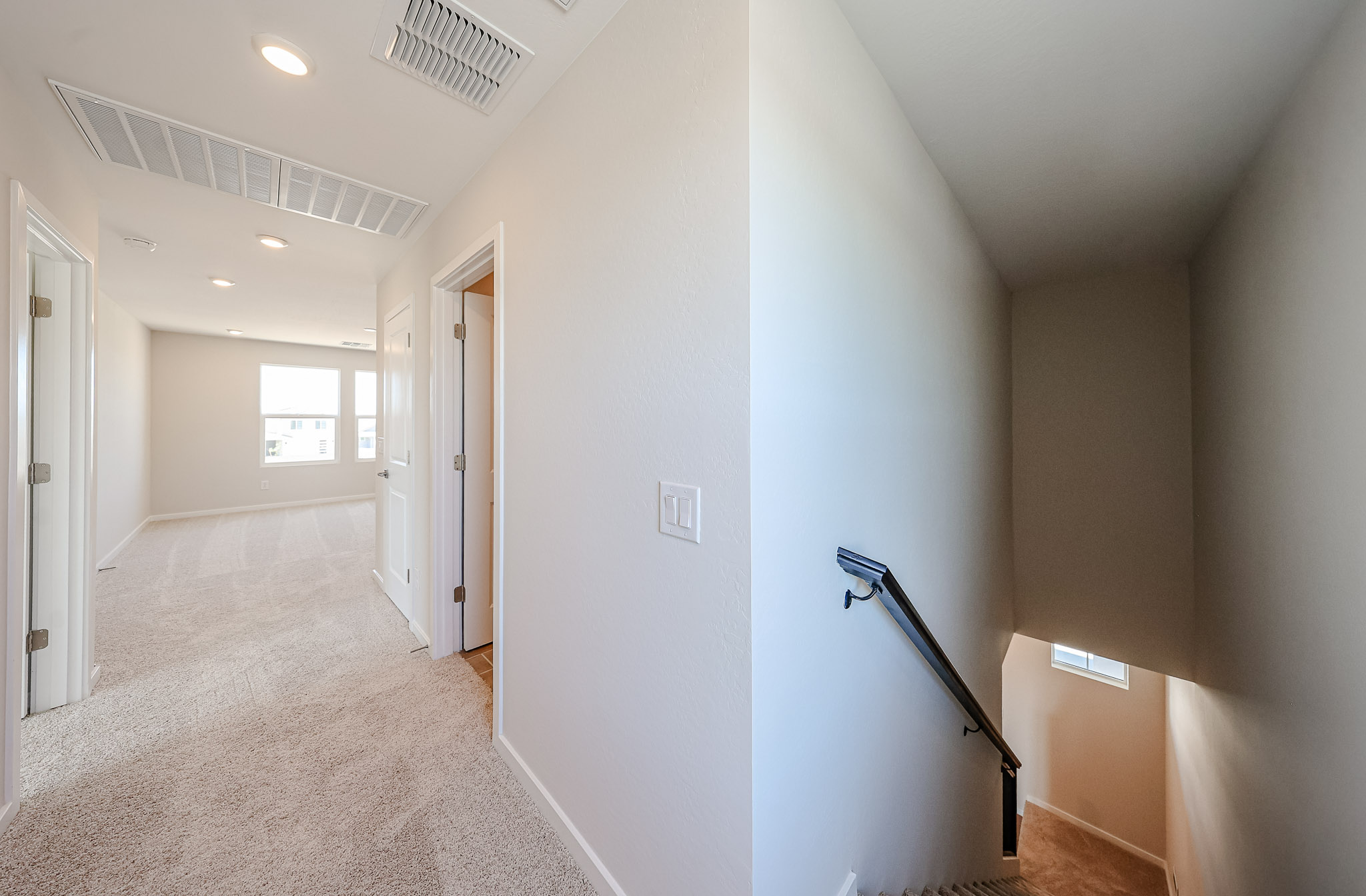 Upstairs hallway with beige carpet, white walls, three doors on the left, and a staircase with a black handrail leading down near a small window.