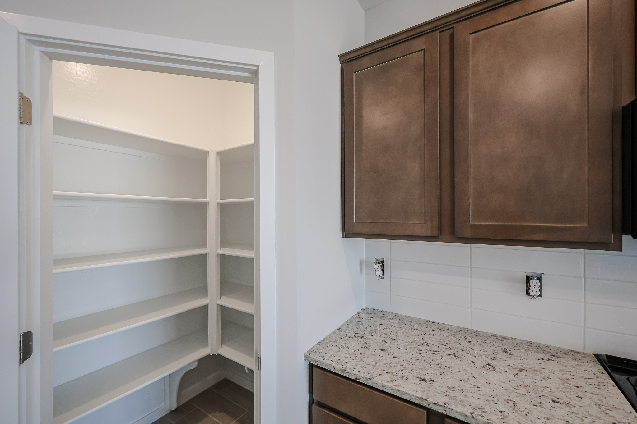 A kitchen with brown cabinets and granite countertop next to an open door revealing empty white shelves in a pantry.