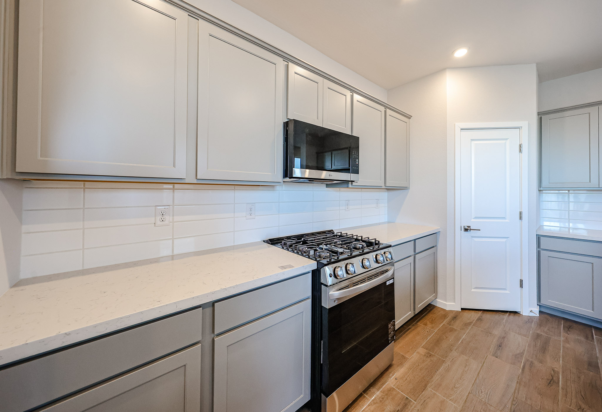 Modern kitchen with grey cabinets, a stainless steel gas stove, black microwave, white countertops, white tile backsplash, and wood flooring.