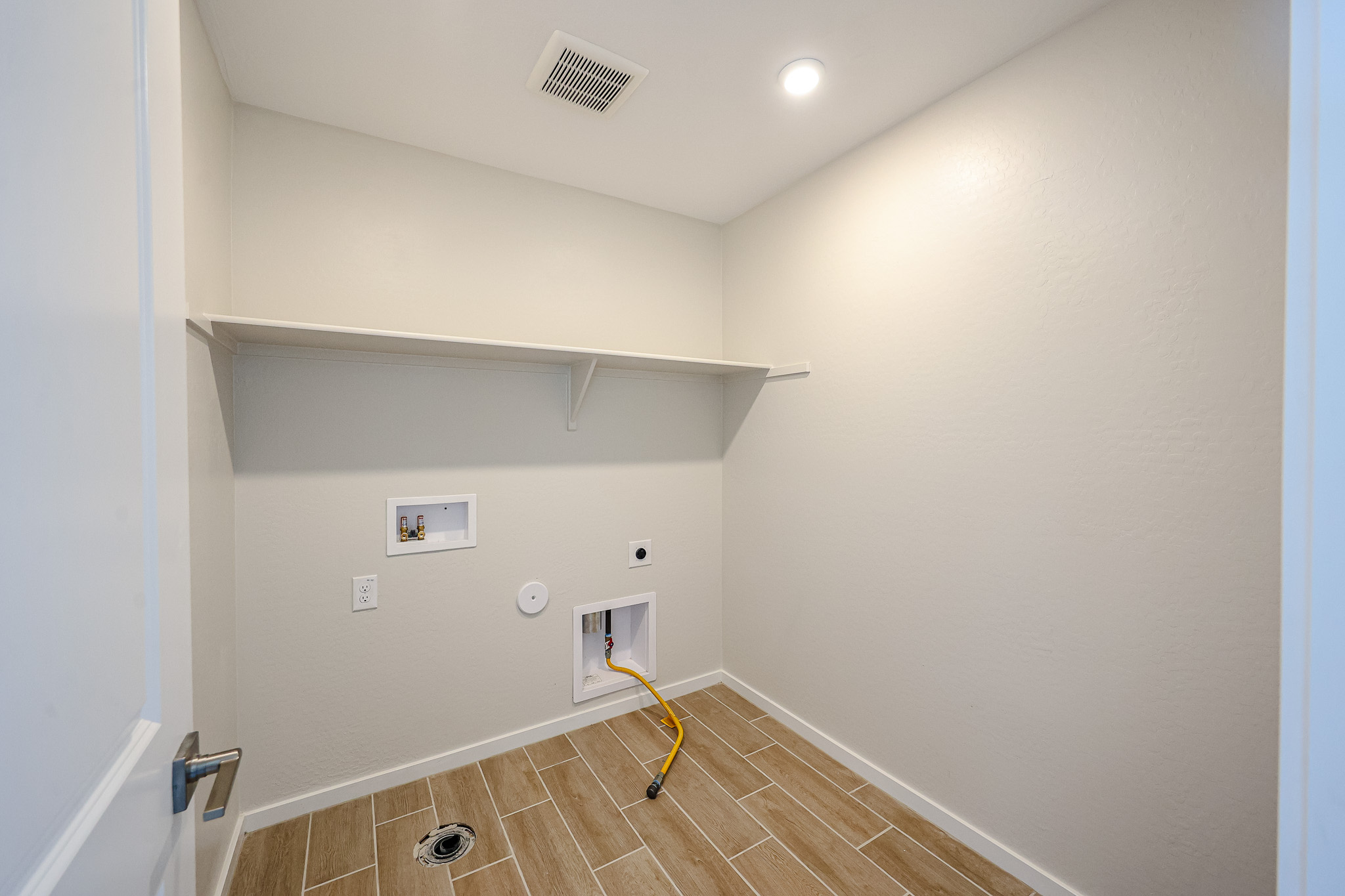 Empty laundry room with beige walls, wood-look tile floor, a wall shelf, hookup outlets for washer and dryer, and ceiling light.