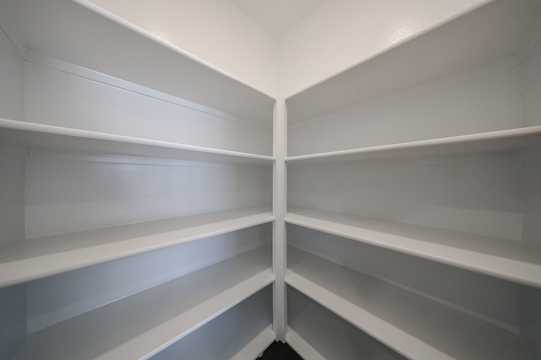 Empty walk-in pantry with white built-in shelves on two walls, extending from floor to ceiling, under bright lighting.
