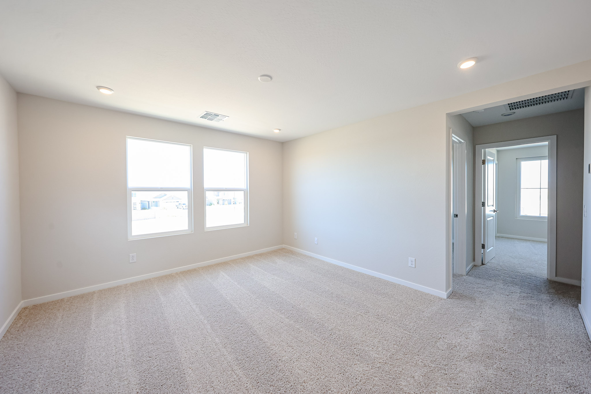Empty, carpeted room with two large windows, light-colored walls, recessed ceiling lights, and a hallway leading to other rooms.