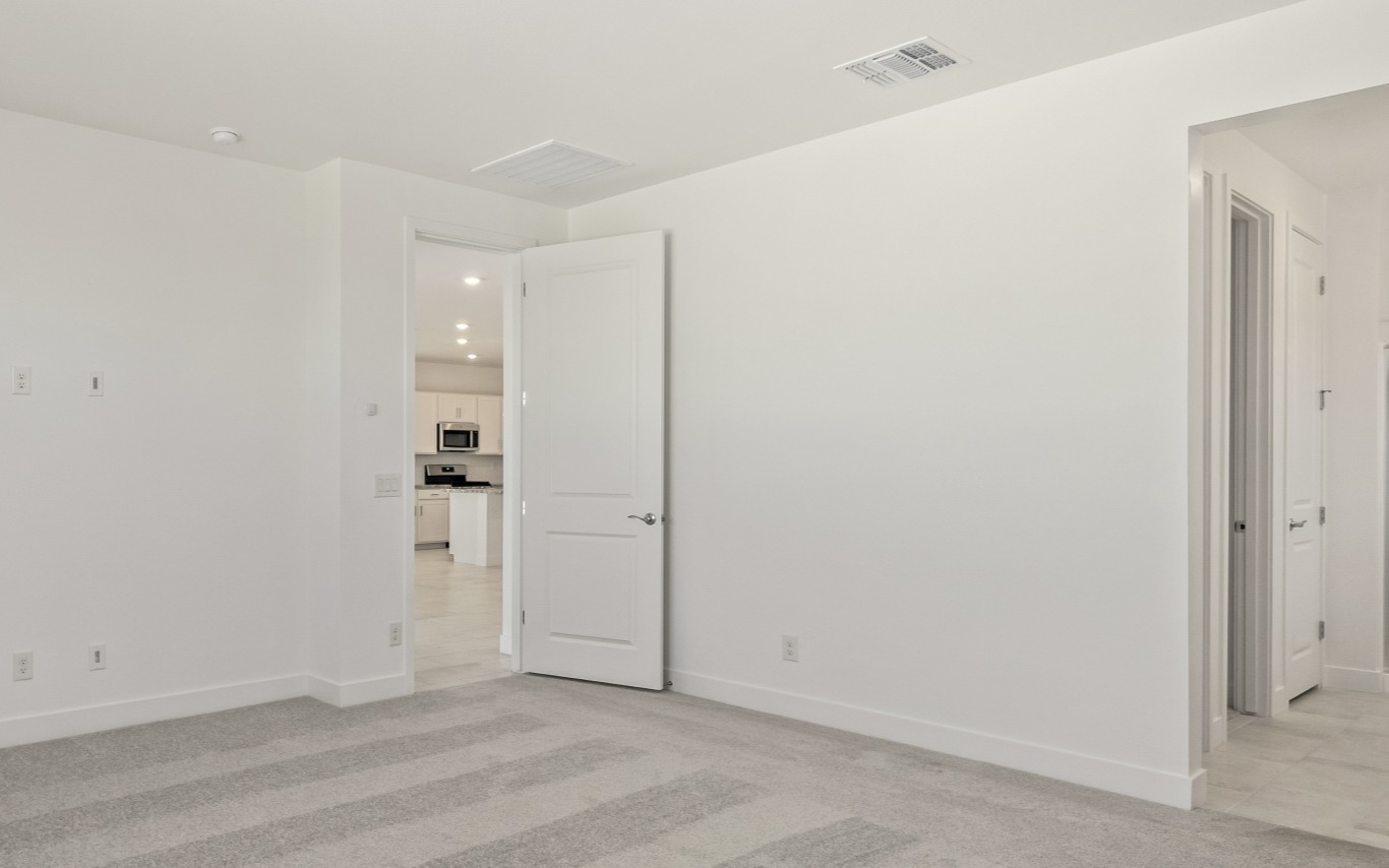 An empty room with light gray carpet and white walls, featuring an open doorway revealing a kitchen in the background and a closed doorway on the right.