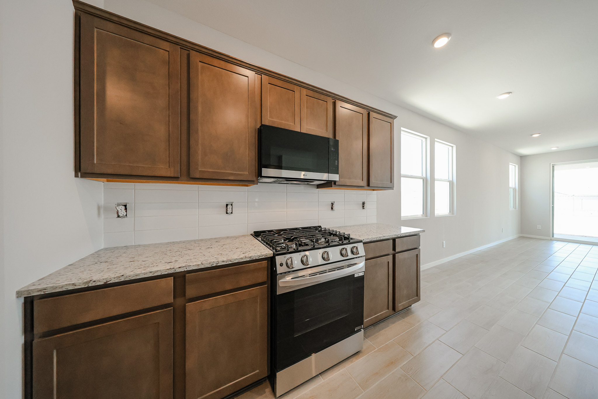 Modern kitchen with brown cabinets, granite countertops, a stainless steel gas stove, and a black microwave, set against a white tile backsplash and a tiled floor.