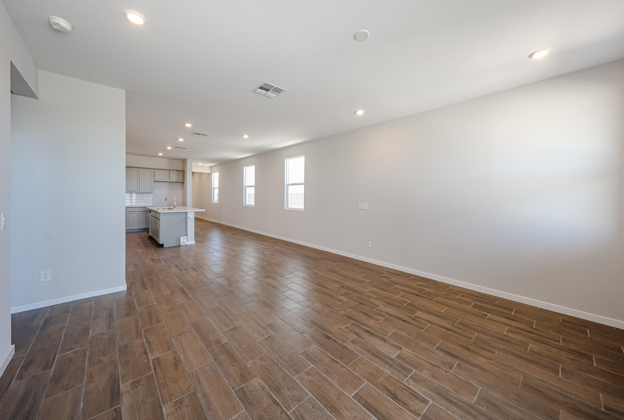 Open-plan room with wood-look tile flooring, white walls, recessed lighting, and a modern kitchen visible in the background. Multiple windows line the right wall.