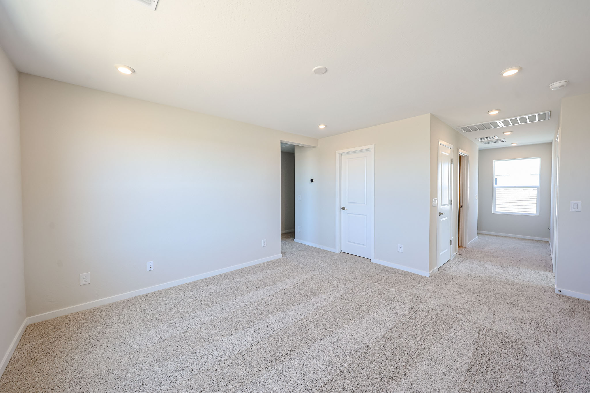 Empty, carpeted room with recessed lighting, beige walls, and white doors leading to a hallway with a window at the end.