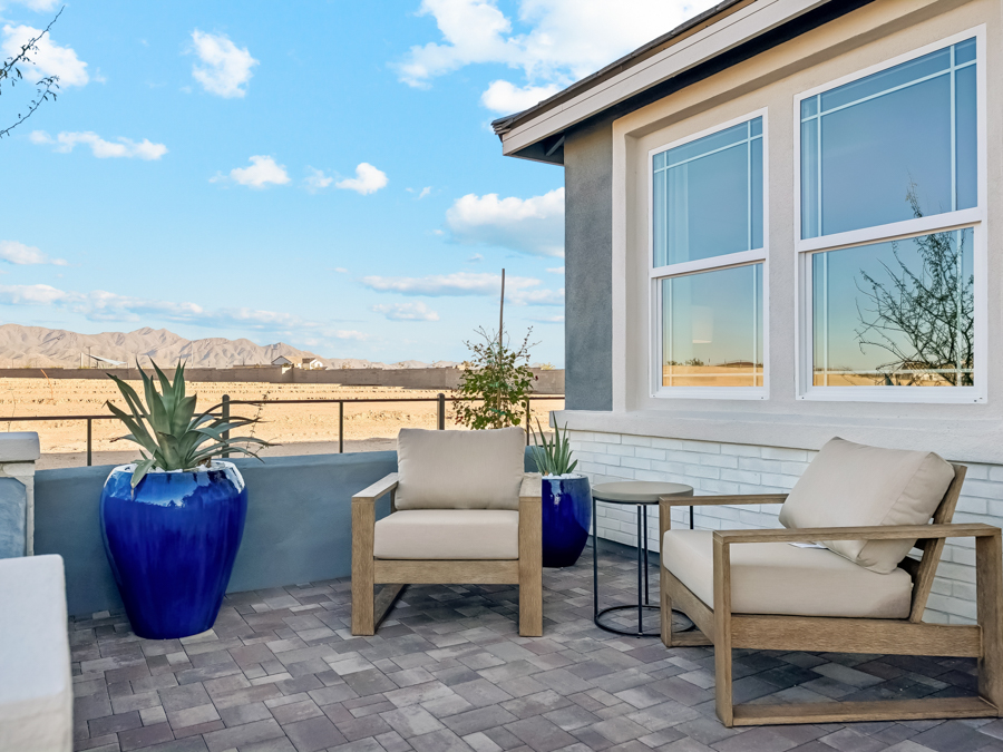 Modern patio with two cushioned chairs, a small table, and two large blue planters. A desert landscape and mountains are visible in the background under a partly cloudy sky.