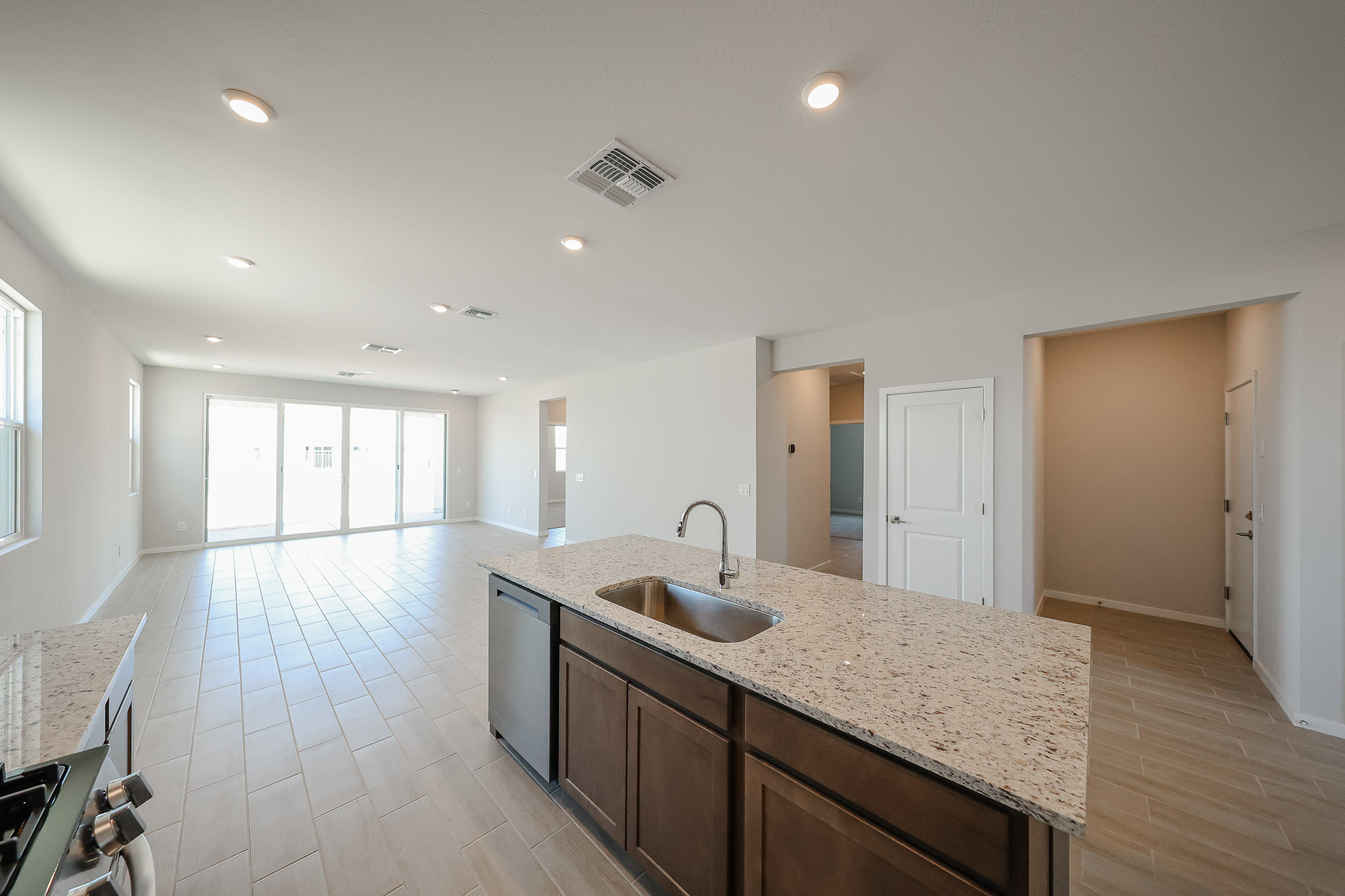 Modern kitchen with granite island, sink, and dark cabinets, opening into a spacious, bright living area with tile flooring and large sliding glass doors.