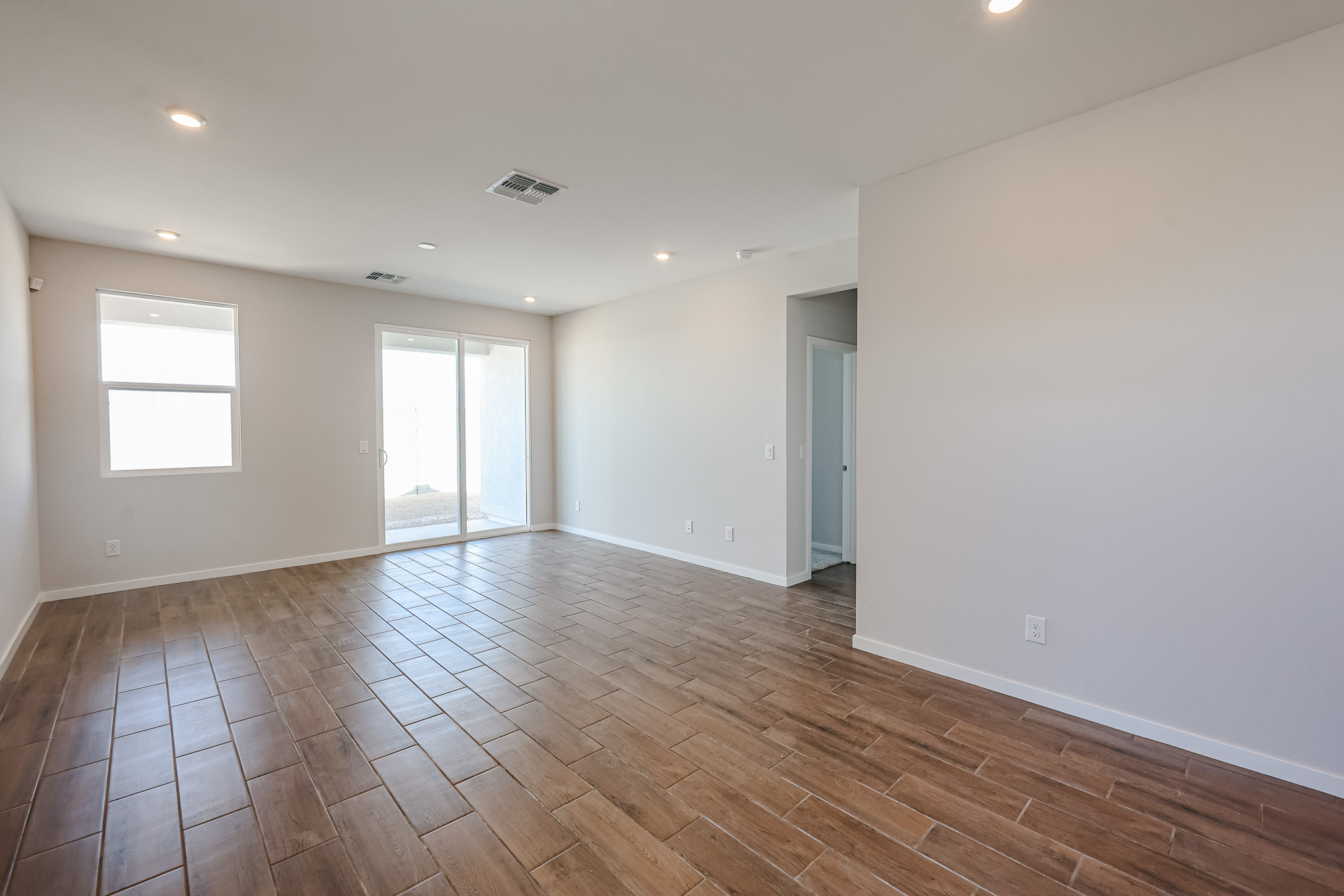 Empty room with brown tile flooring, white walls, recessed lighting, a large window, sliding glass doors, and access to a hallway.