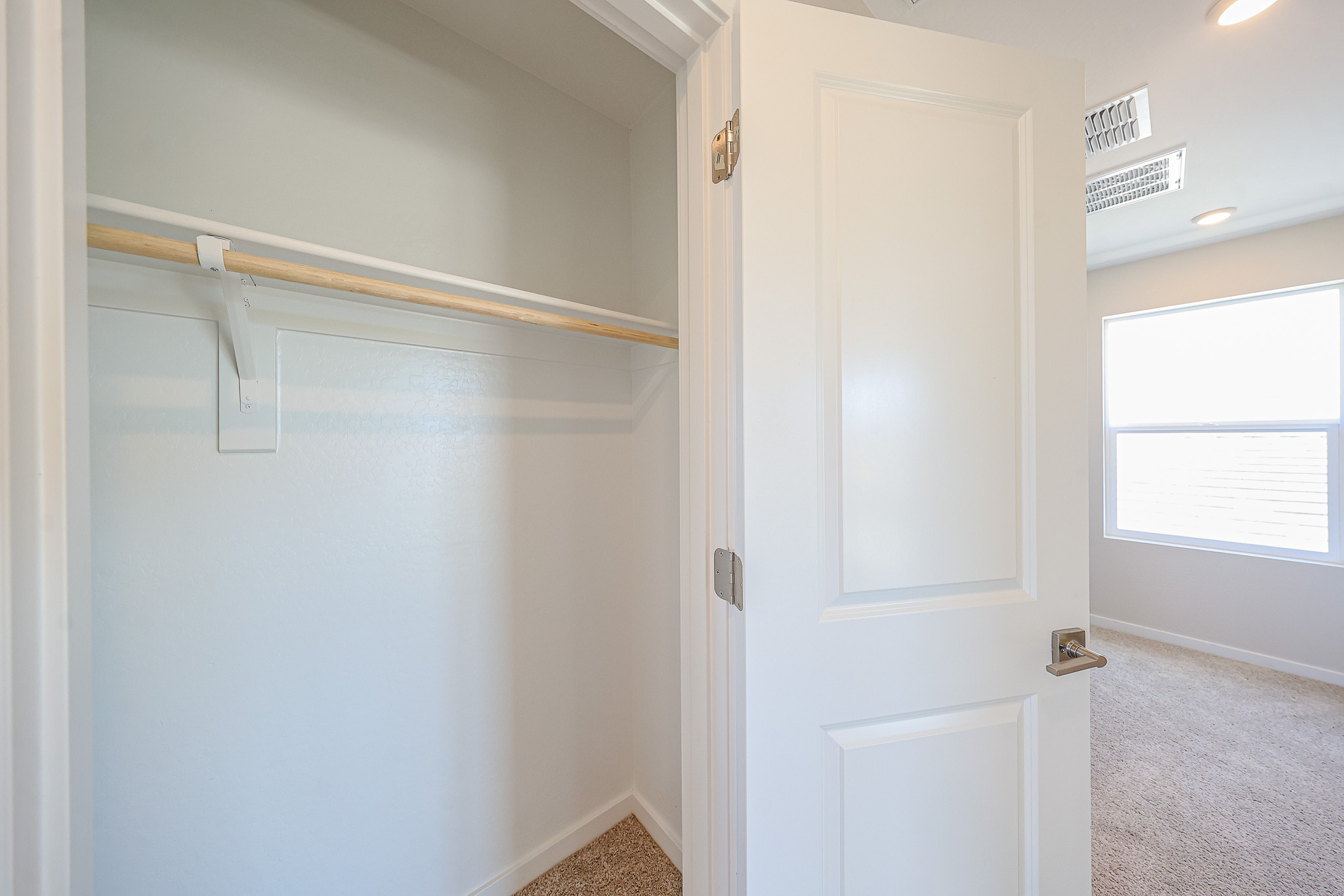 Open white closet door reveals an empty closet with a single wooden rod and shelf, beige carpet flooring, and white walls in a well-lit room.