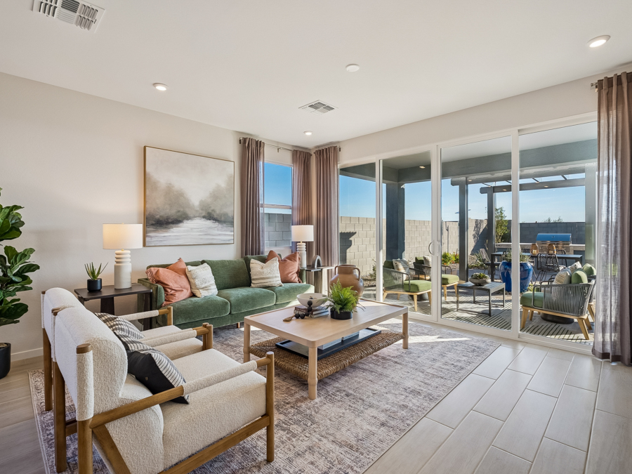 Modern living room with green sofa, two armchairs, coffee table, large window, and sliding glass doors opening to a patio with outdoor seating. Neutral colors and contemporary decor.